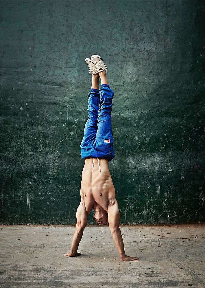 A shirtless adult male performs a handstand outdoors wearing blue pants and white sneakers, showcasing strength and balance.