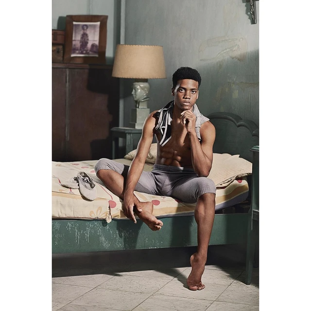 Young adult male sitting on a bed, shirtless with grey athletic leggings and towel around his neck, with cream, green, and grey colour palette.