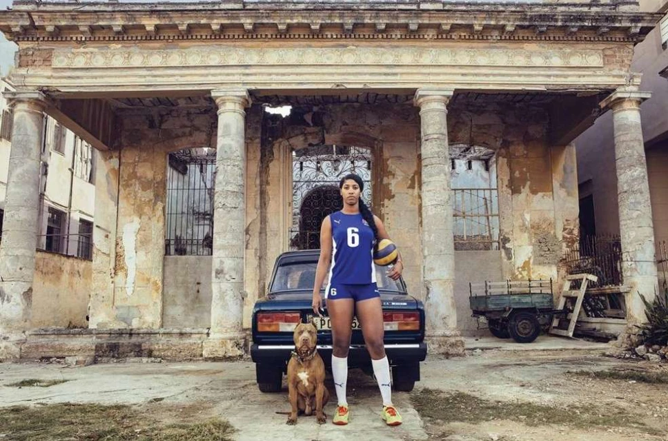 A female volleyball player poses with a dog and a volleyball in front of a vintage car and weathered building in an urban setting.