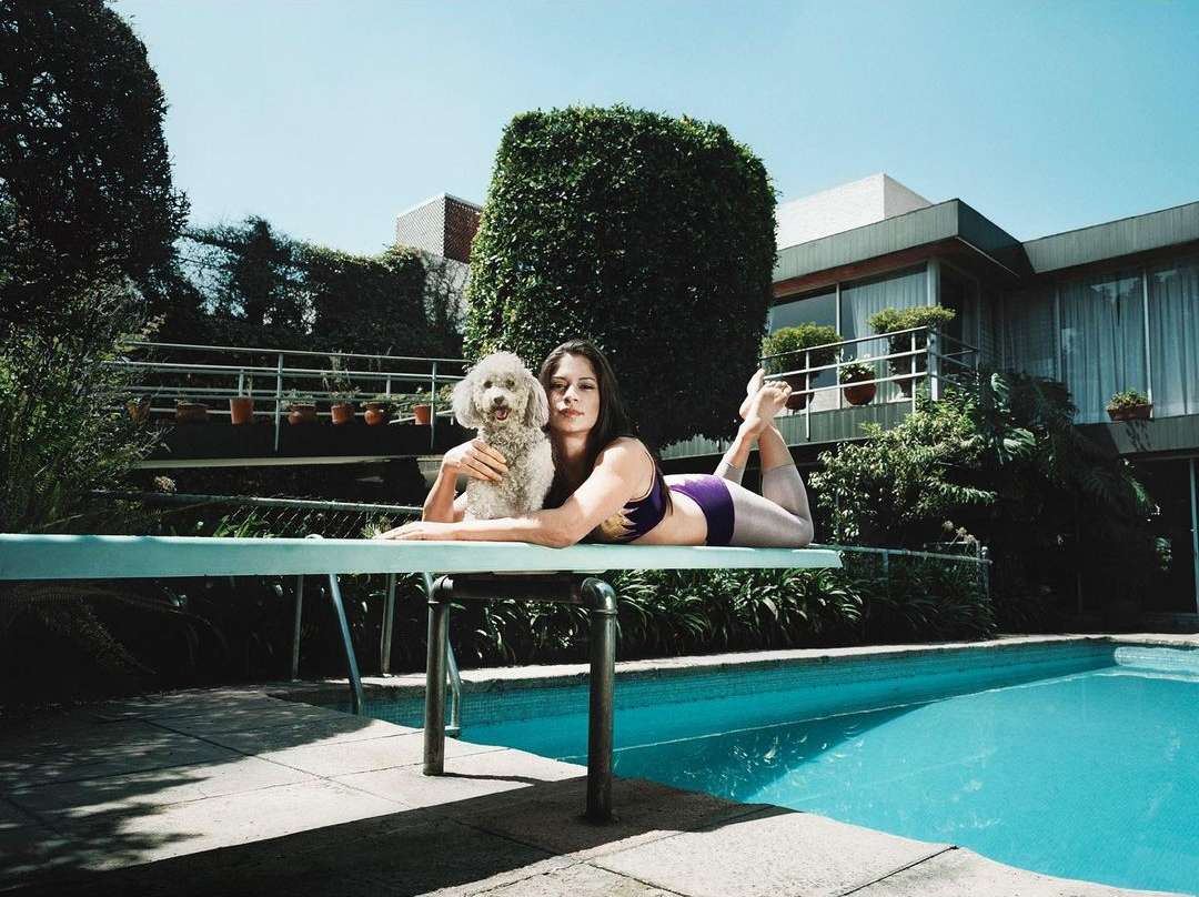 Woman in swimsuit lies on diving board with poodle next to blue swimming pool, green bushes, and modern house on sunny day.