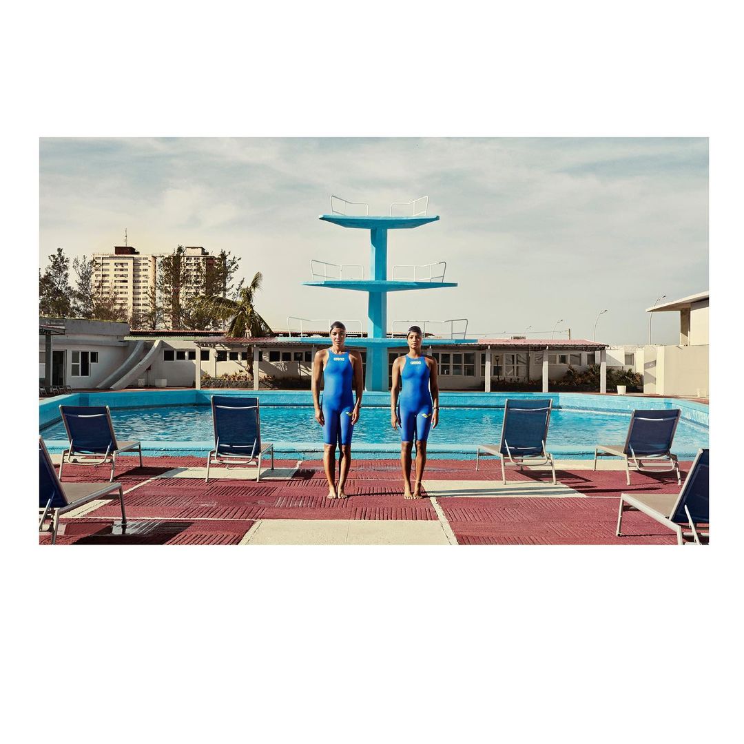 Two female swimmers in blue swimsuits stand poolside near a blue diving platform and pool, under a bright sky with buildings visible.