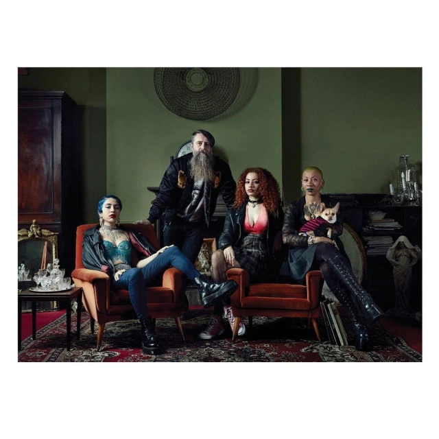 Group portrait of four alternative-styled adults with a small dog indoors in a room with vintage decor and olive green walls.