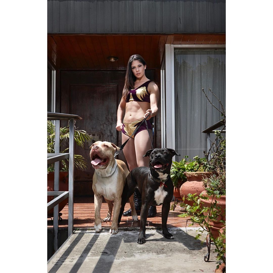Female wrestler poses with two pit bull dogs in front of a house, wearing a purple and gold wrestling outfit with brown hair.