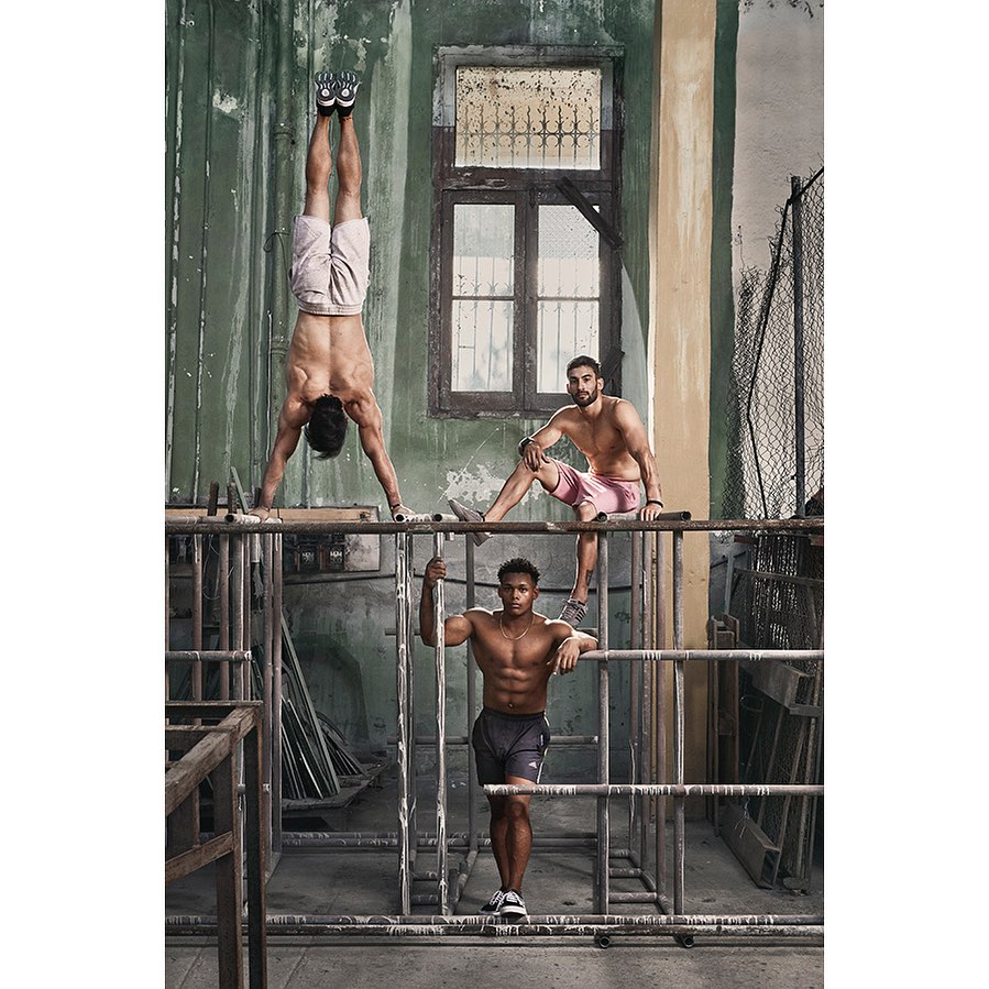 Three athletic men strike poses on a metal structure inside an industrial space. One man is doing a handstand, showcasing strength and balance.