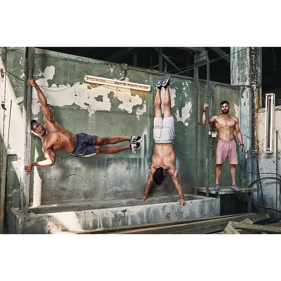 Three muscular men demonstrate advanced calisthenics poses against a weathered concrete wall in an industrial-style setting.
