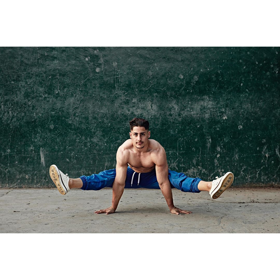Bare-chested man in blue sweatpants performing handstand split outdoors on concrete against a textured dark green wall, fitness.