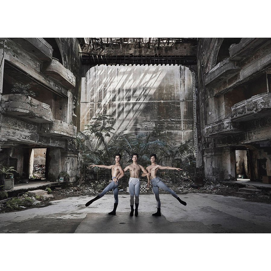 Three male ballet dancers strike a pose in a dilapidated theatre with peeling walls and tropical greenery in the background.