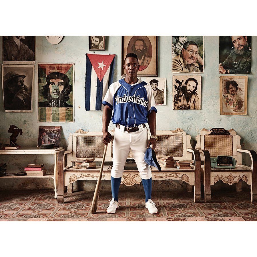 Full shot of a baseball player in uniform, holding a bat and hat, posed indoors against a wall with Cuban flags and portraits.