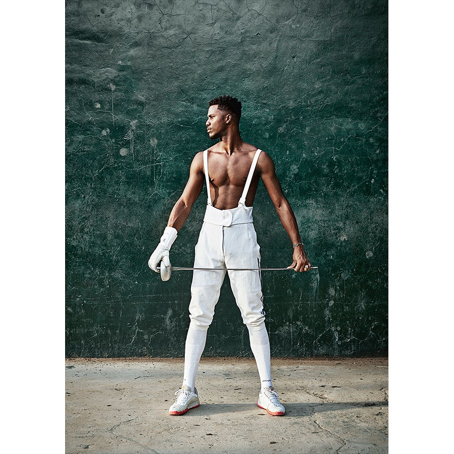 Athletic man in white fencing uniform, holding a foil, posing in front of a weathered green wall, showcasing fitness and classical sport.
