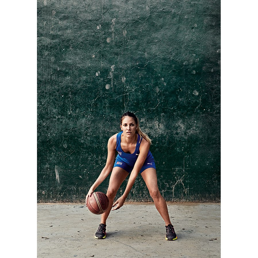 Athletic woman in blue sportswear dribbling a basketball on a grey concrete court against a green, textured wall. Medium shot.