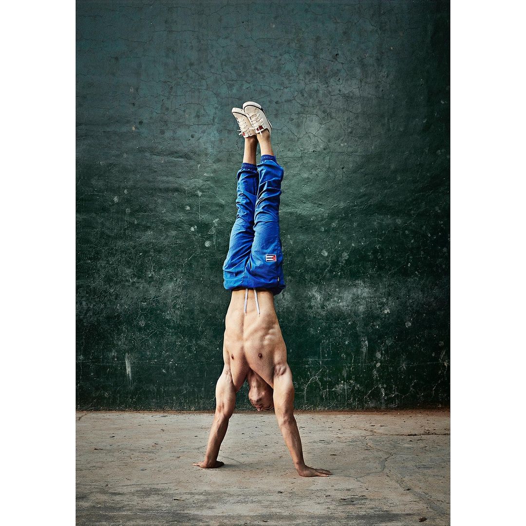Man in blue pants and shoes does a handstand against a dark green textured wall on a concrete surface in a studio.