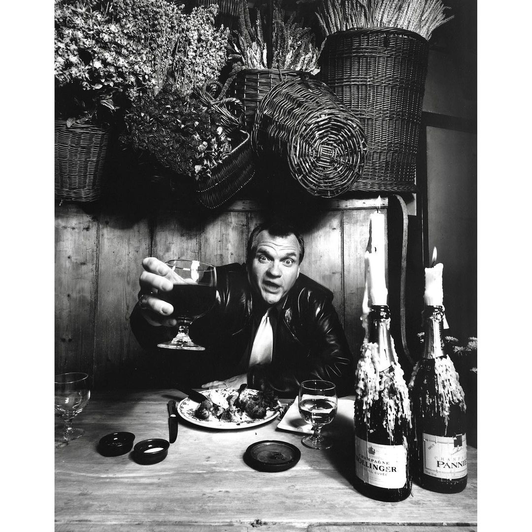 Dramatic black and white portrait of an adult man raising a wine glass at a dining table, illuminated by candlelight, featuring floral arrangement.