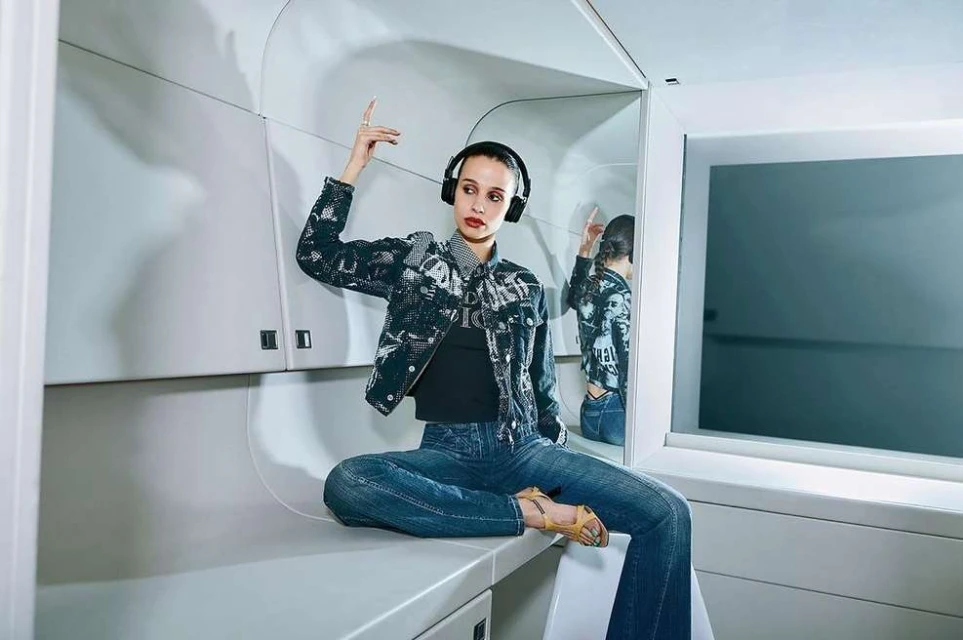 Stylish young woman with headphones poses indoors wearing patterned denim jacket, jeans, and open-toe heels, reflected in a mirror.