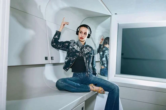 Stylish young woman with headphones poses indoors wearing patterned denim jacket, jeans, and open-toe heels, reflected in a mirror.