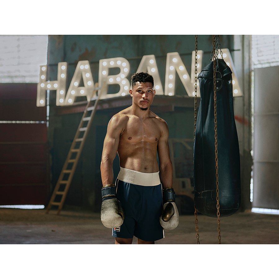 Shirtless boxer wearing gloves and boxing shorts stands in a boxing gym with a punching bag and 'Habana' sign in the background.