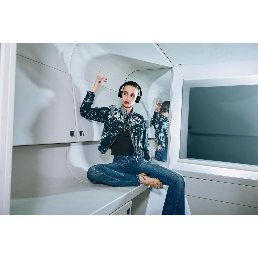 A young woman sits cross-legged on a white surface, wearing headphones, a black top, a jacket with graphic prints, and blue jeans. She is in an enclosed area with white walls and a mirror.