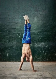 Man performing a handstand against a weathered green wall wearing blue sweatpants and sneakers.