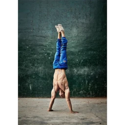 Shirtless man performing a handstand against a weathered dark green wall wearing blue trousers and light-colored sneakers
