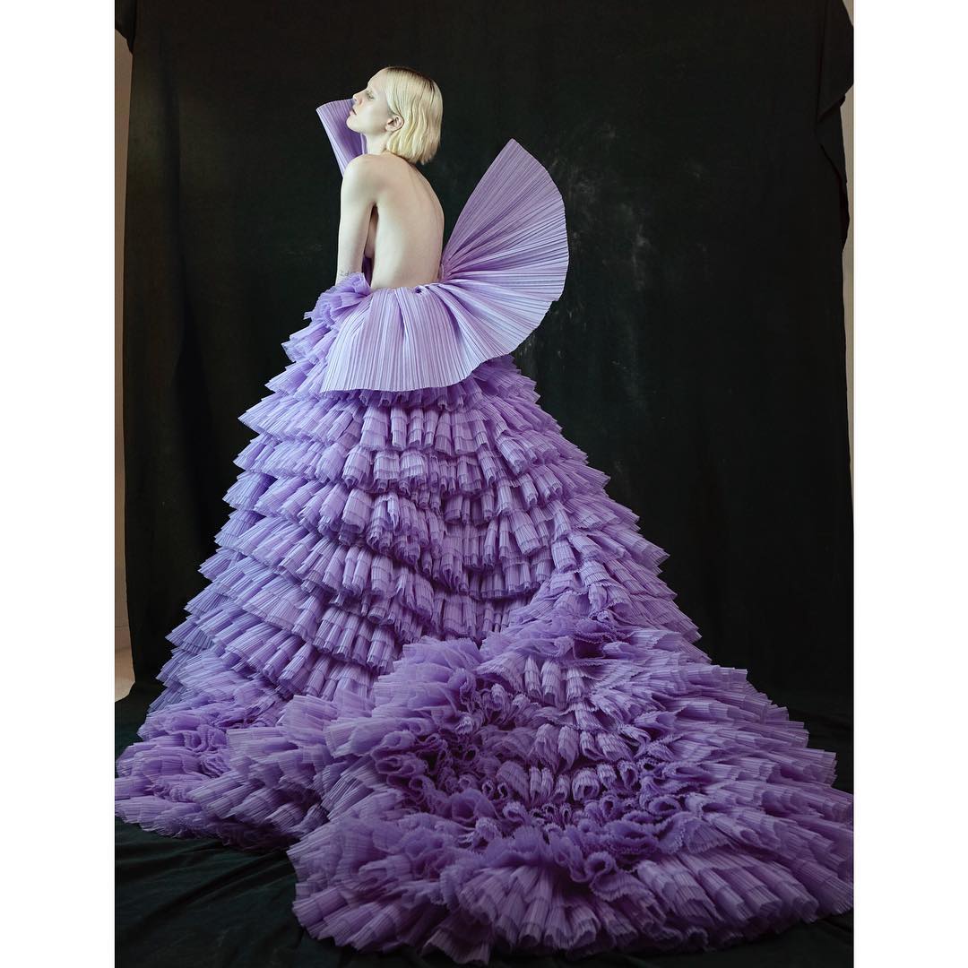 A woman poses in a ruffled, multi-tiered lavender gown against a dark backdrop in a studio fashion shot with a low-angle perspective.
