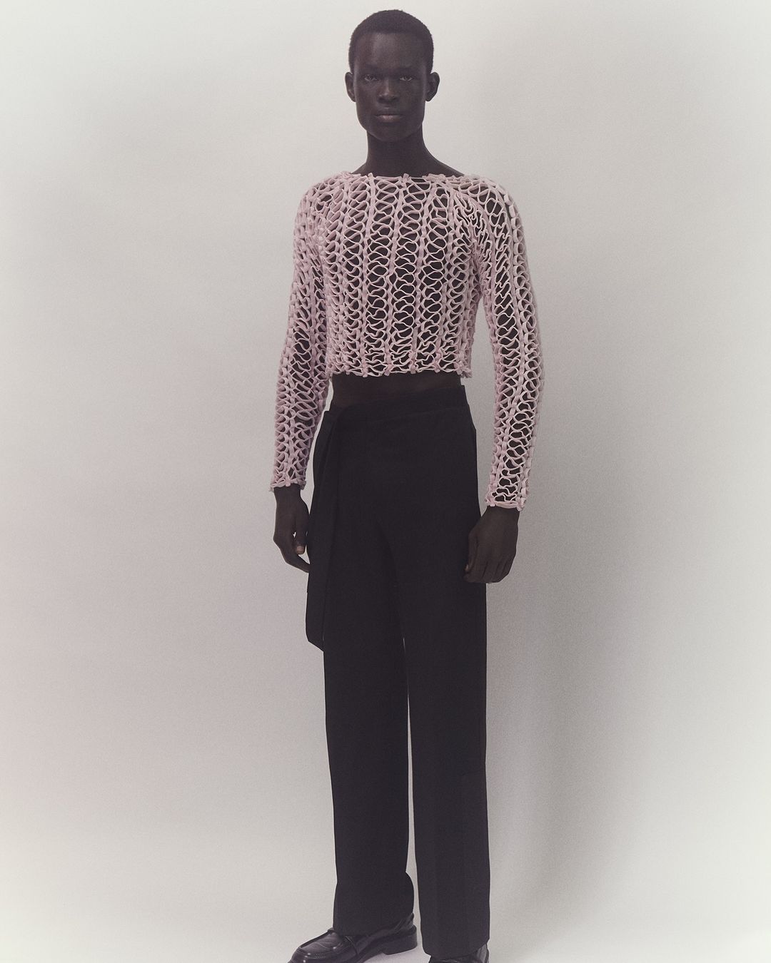 Full-length studio shot of adult male fashion model wearing a pink mesh top and black pants on a plain white background, modern fashion.