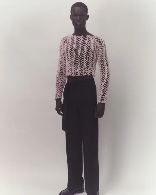 Full-length studio shot of adult male fashion model wearing a pink mesh top and black pants on a plain white background, modern fashion.