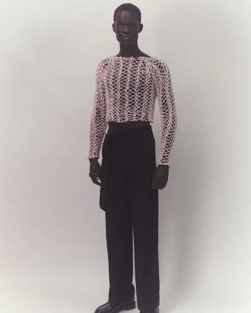 Full-length studio shot of adult male fashion model wearing a pink mesh top and black pants on a plain white background, modern fashion.