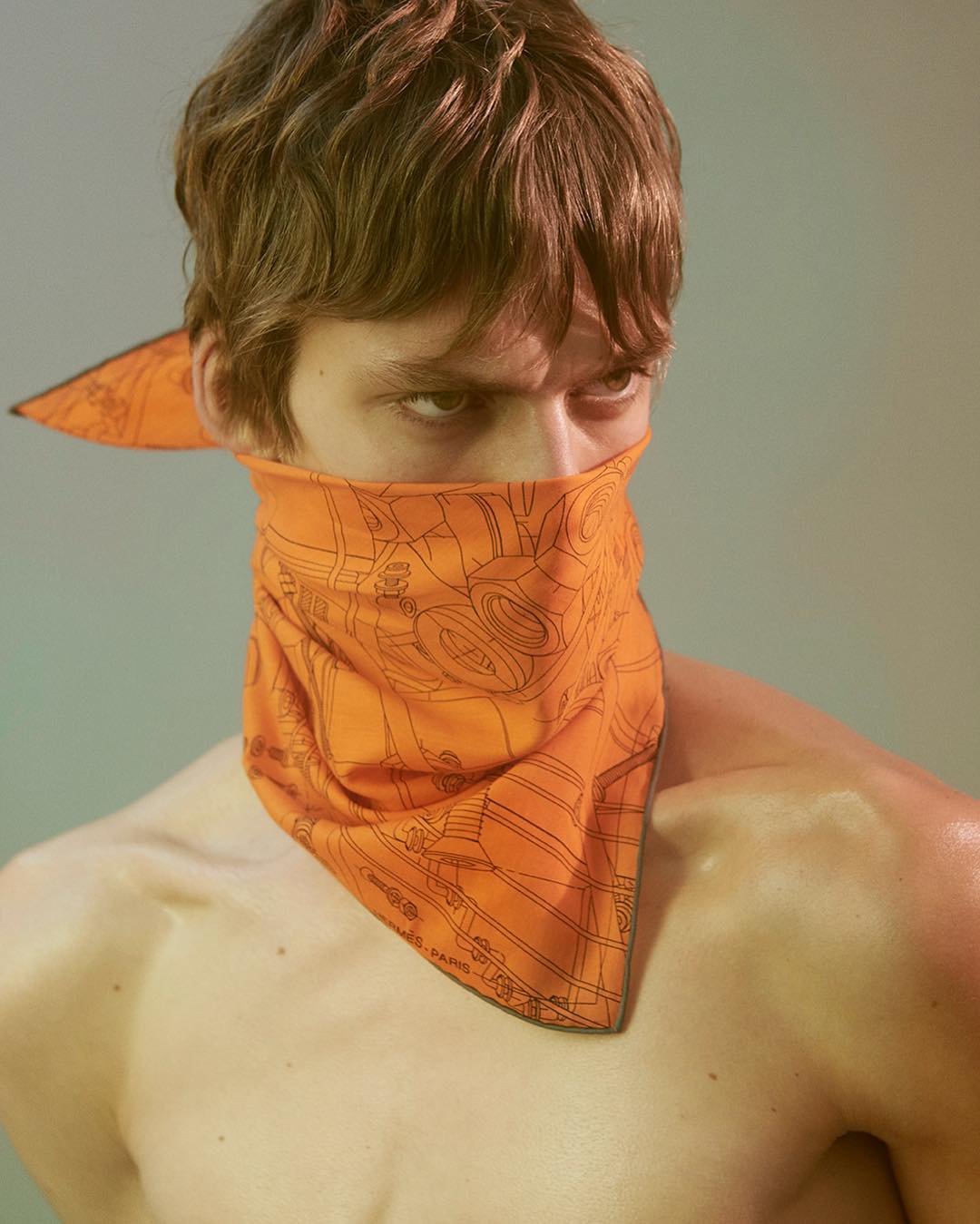 Portrait of a young adult man with brown hair wearing an orange patterned kerchief covering the lower half of his face indoors.