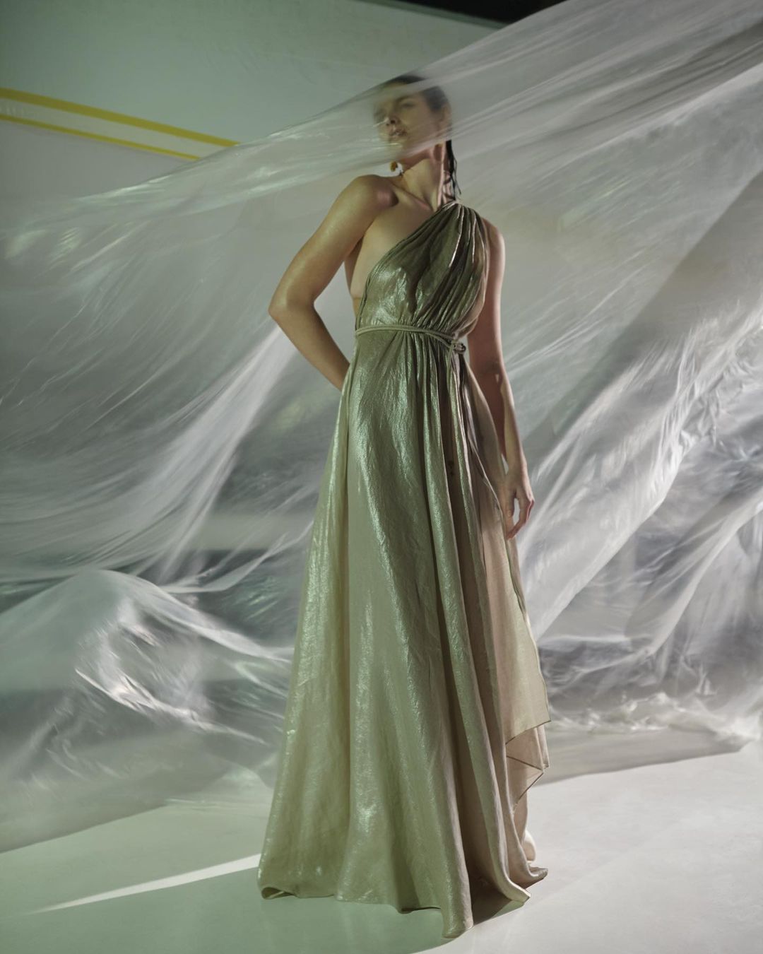 Elegant adult woman poses in shimmering gold, one-shoulder gown with draped detail, set against a flowing sheer backdrop and soft studio light.