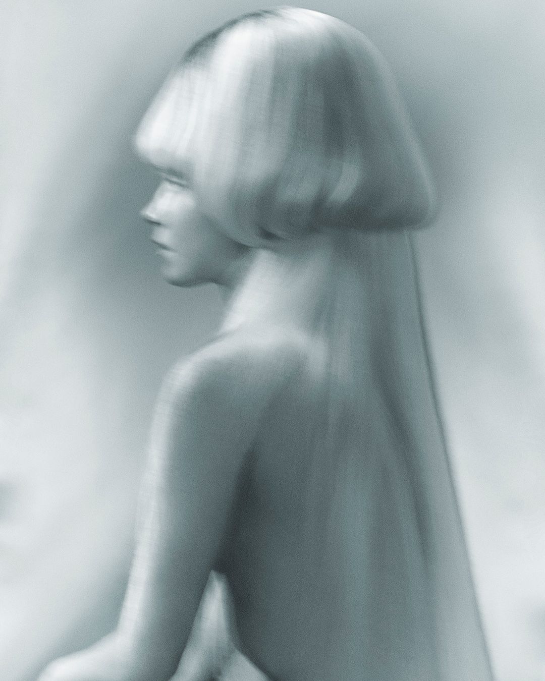 Monochromatic close-up of a woman with a blonde bob hairstyle in a profile pose with her back visible against a blurred background.