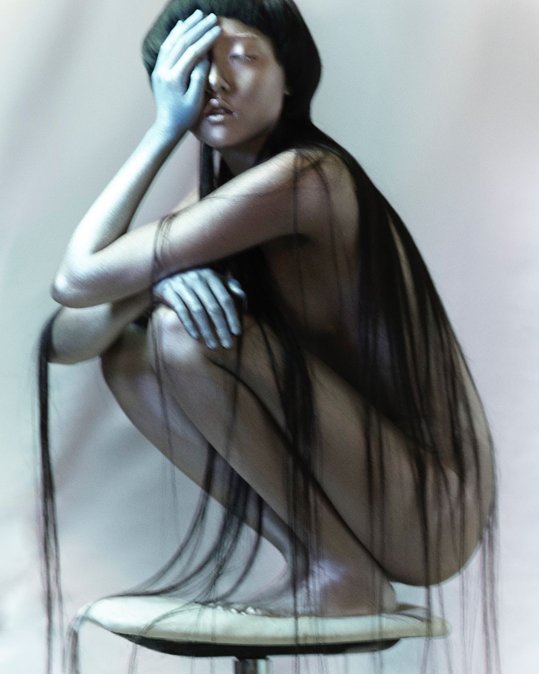 Adult female crouching on a white stool with face partially covered by hand, long black hair falling over stool. Silver body paint visible.