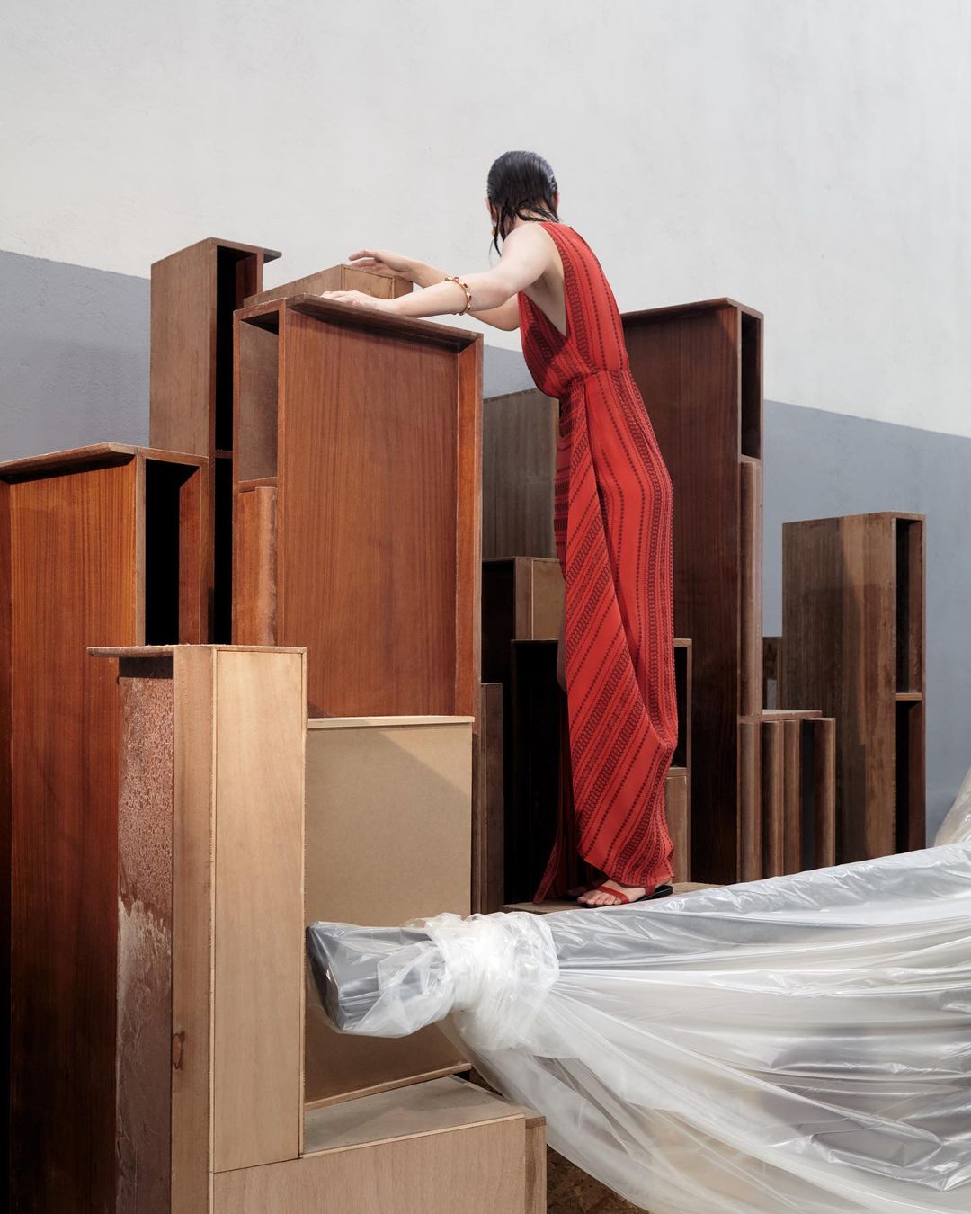 A woman in a red dress stands among stacked wood boxes, creating an urban landscape effect, with a neutral wall backdrop in a studio setting.