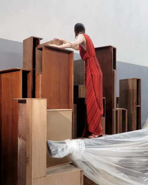 A woman in a red dress stands among stacked wood boxes, creating an urban landscape effect, with a neutral wall backdrop in a studio setting.