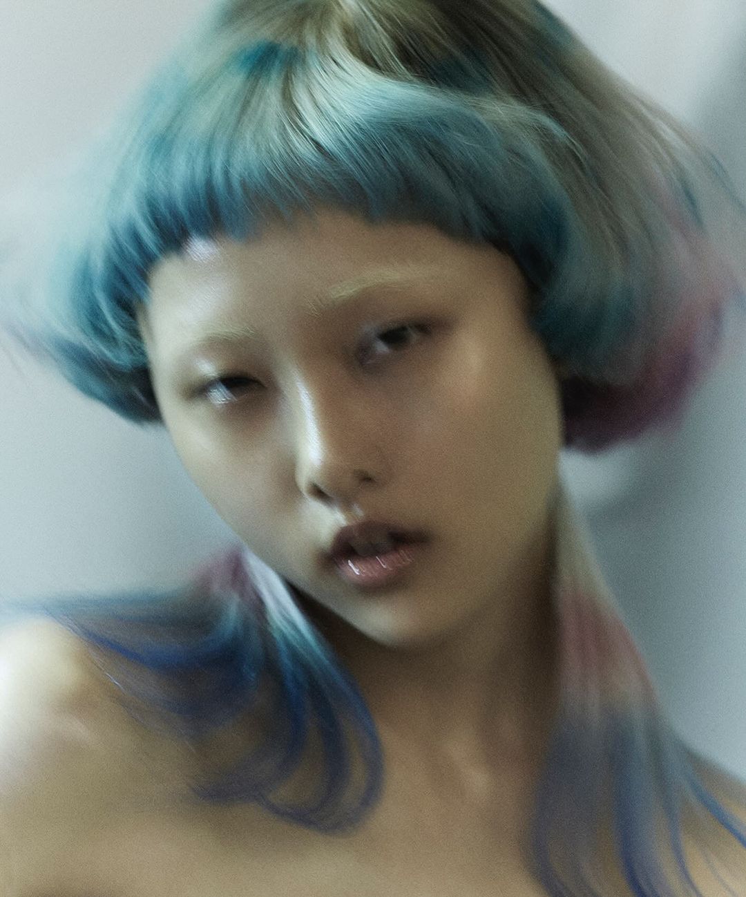 Portrait of an Asian woman with blue and pink dyed short hair, glossy skin, and light make-up, posing indoors with blurred background.