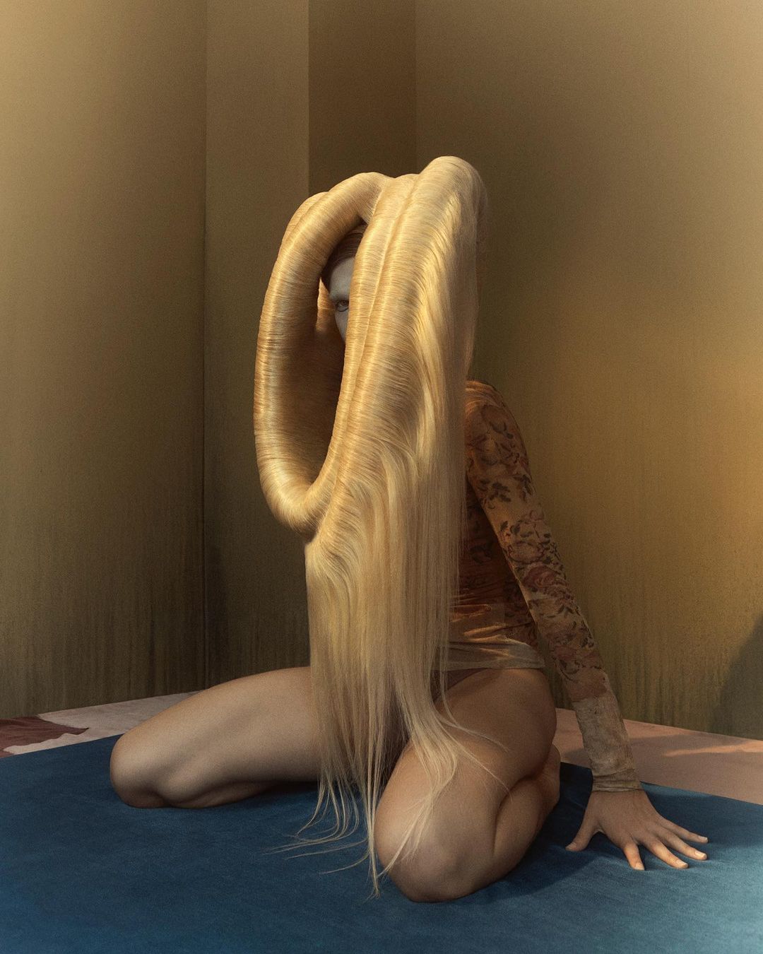 Woman kneels in a beige space, adorned in a light brown lace bodysuit, long blonde hair styled into a looped shape, illuminated by studio lighting.