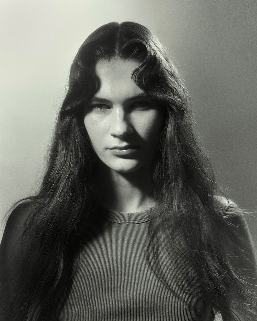 Monochrome portrait of a young woman with long wavy hair, wearing a ribbed crew-neck top, against a softly lit gradient backdrop.