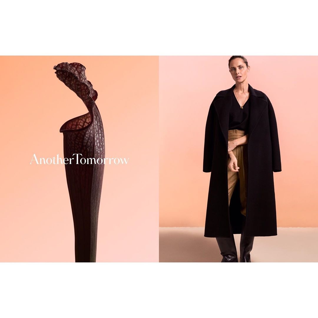 Fashion shot featuring a woman in a black coat and tan pants alongside a dark-colored pitcher plant against a light peach backdrop.