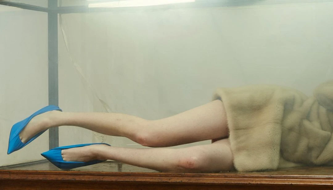 Legs in blue shoes and a tan coat submerged in clear liquid inside of a glass box sit on a wooden table in an indoor setting.