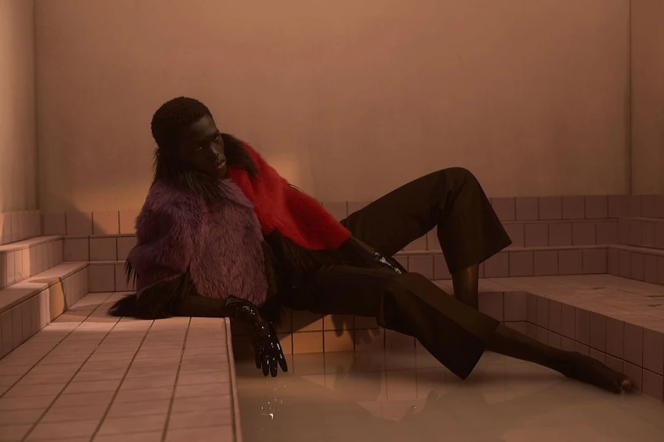 Dark-skinned model reclines in an indoor tiled space with water, wearing a purple faux fur coat, red top, and black pants and gloves.