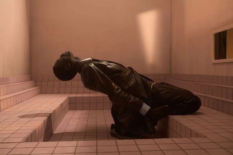 An African-American man in a black suit poses, leaning back in an artistic way in a pink-tiled architectural indoor space with a mirror.