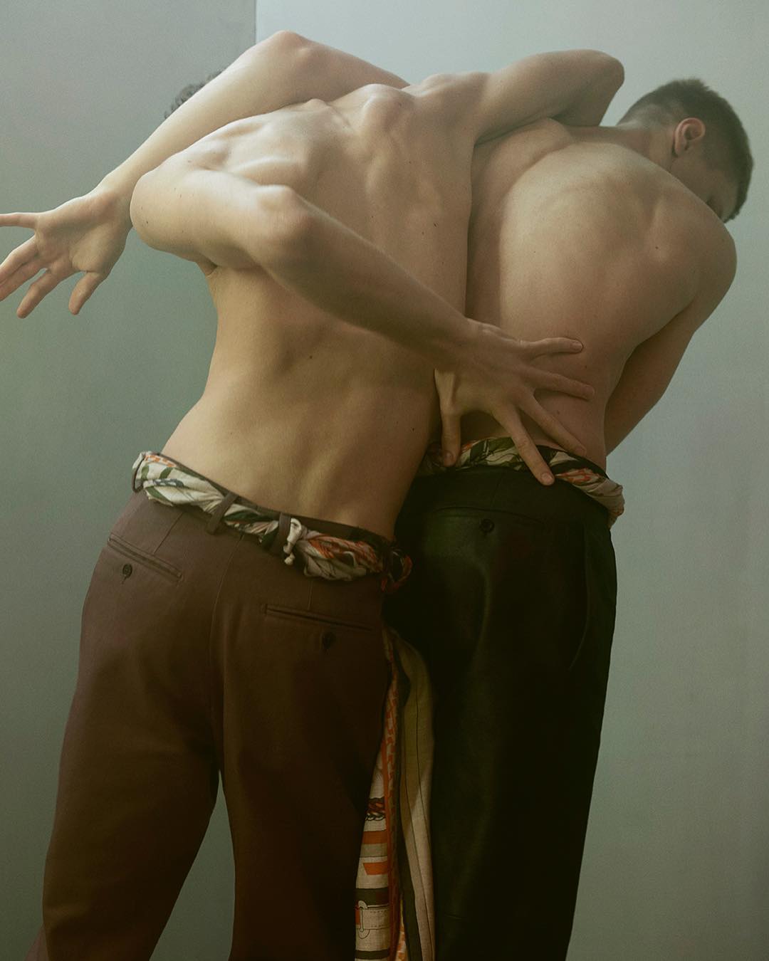 Two shirtless adult men stand closely together with arms intertwined; they are wearing dress pants and colorful patterned cloth belts.