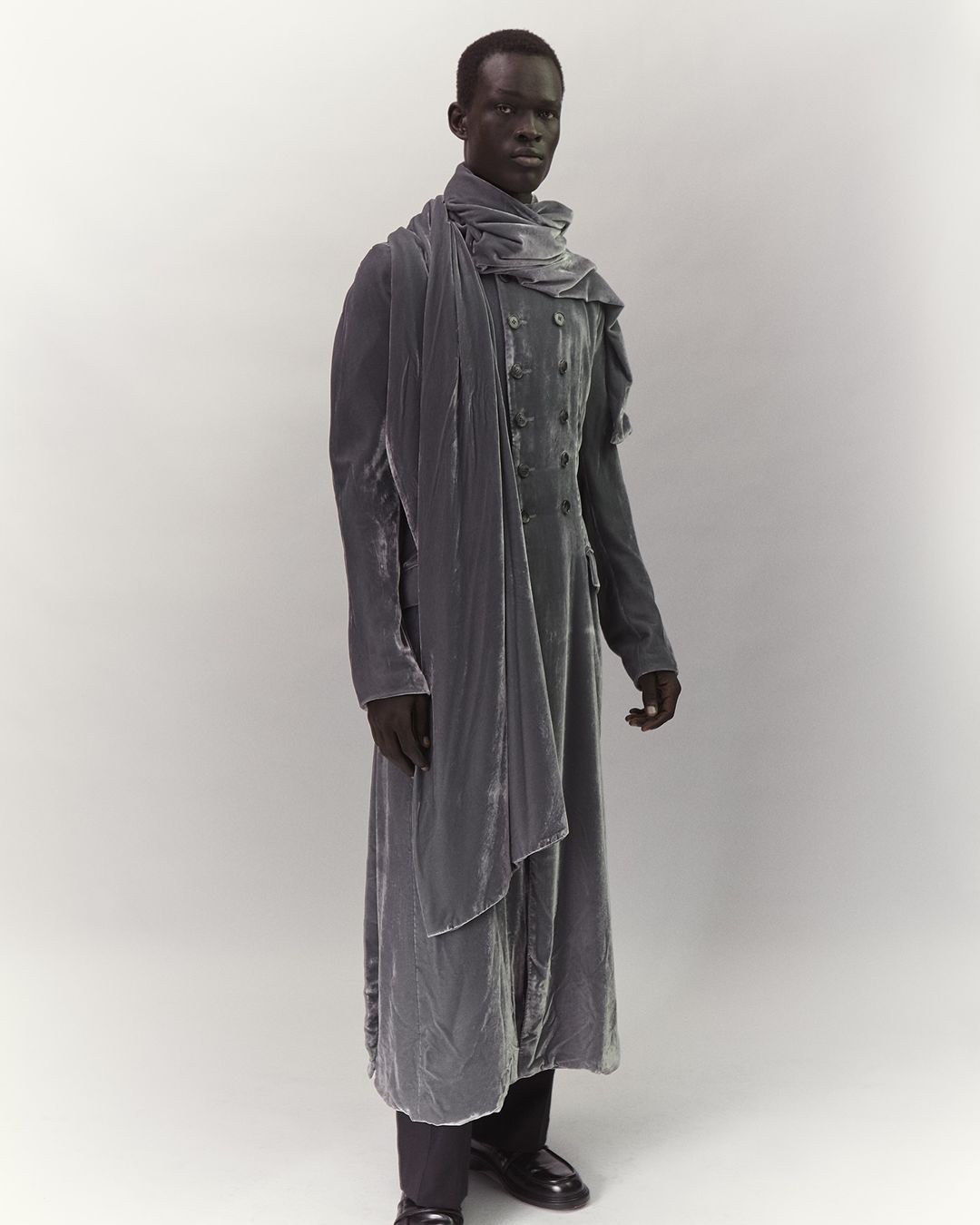 Full-length portrait of a male fashion model wearing a grey velvet coat and scarf in a photo studio, posing in front of a grey backdrop.