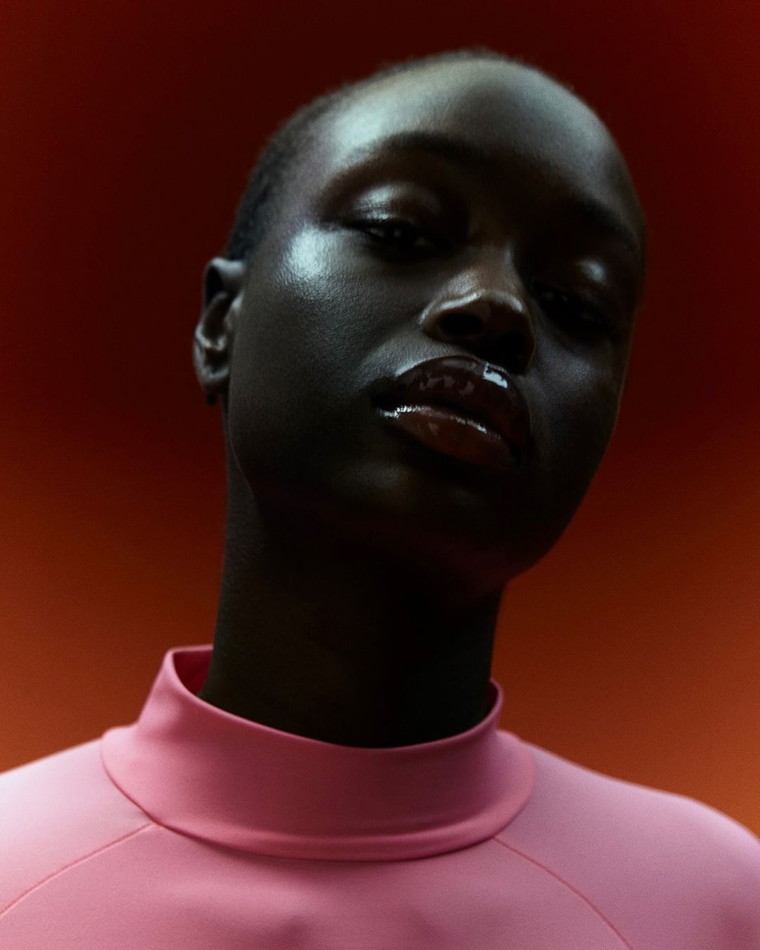 Close-up studio portrait of a Black female model in pink top against a warm orange background, showcasing beauty and fashion aesthetics.
