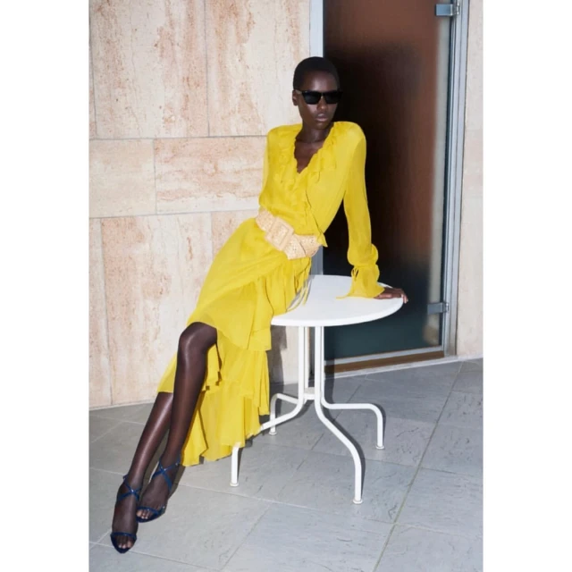A dark-skinned woman models a yellow ruffle dress with long sleeves and a woven belt while sitting on a white table indoors.
