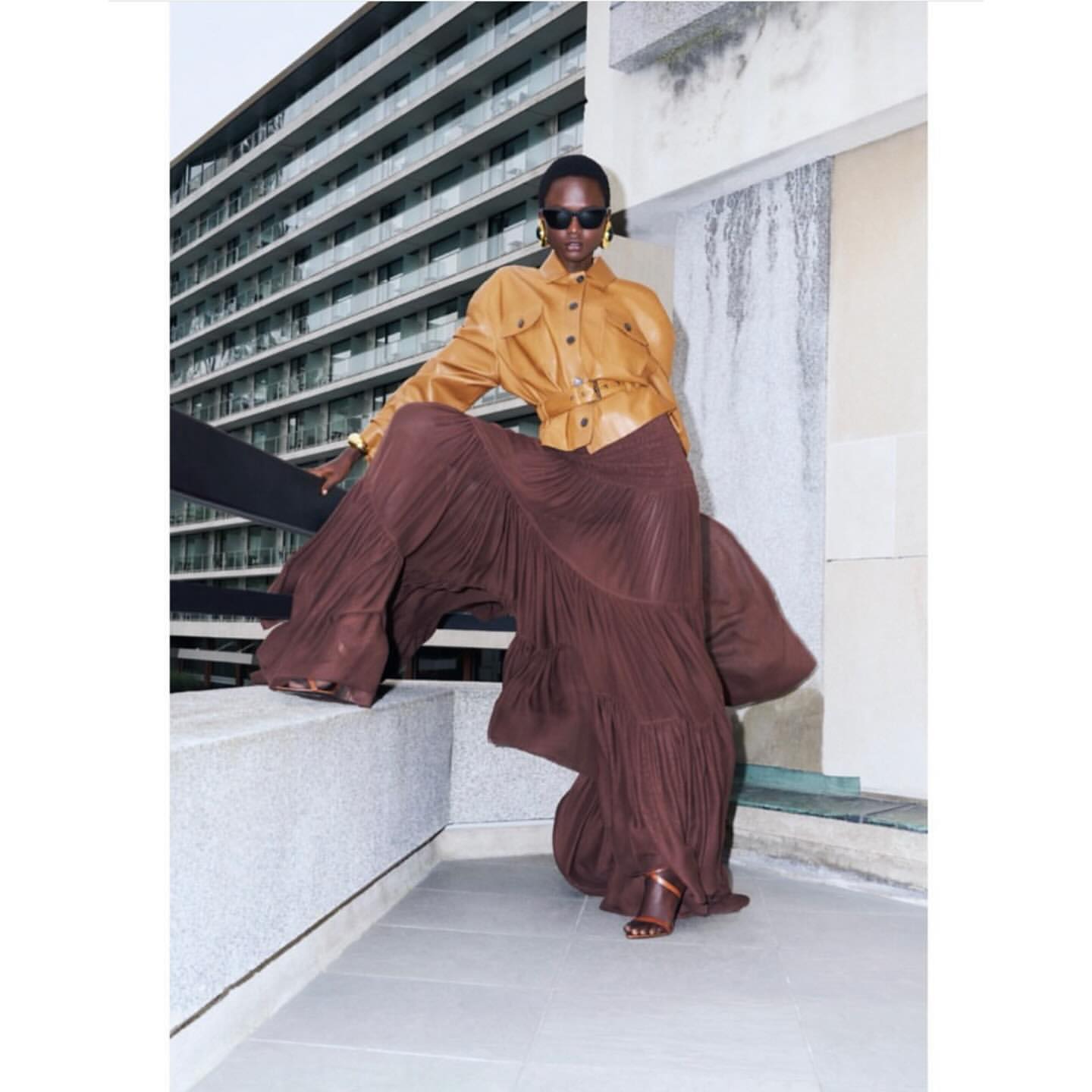 Model wearing dark sunglasses, camel leather jacket, and voluminous brown pleated skirt posing outdoors against a residential building.