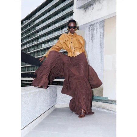 Model wearing dark sunglasses, camel leather jacket, and voluminous brown pleated skirt posing outdoors against a residential building.