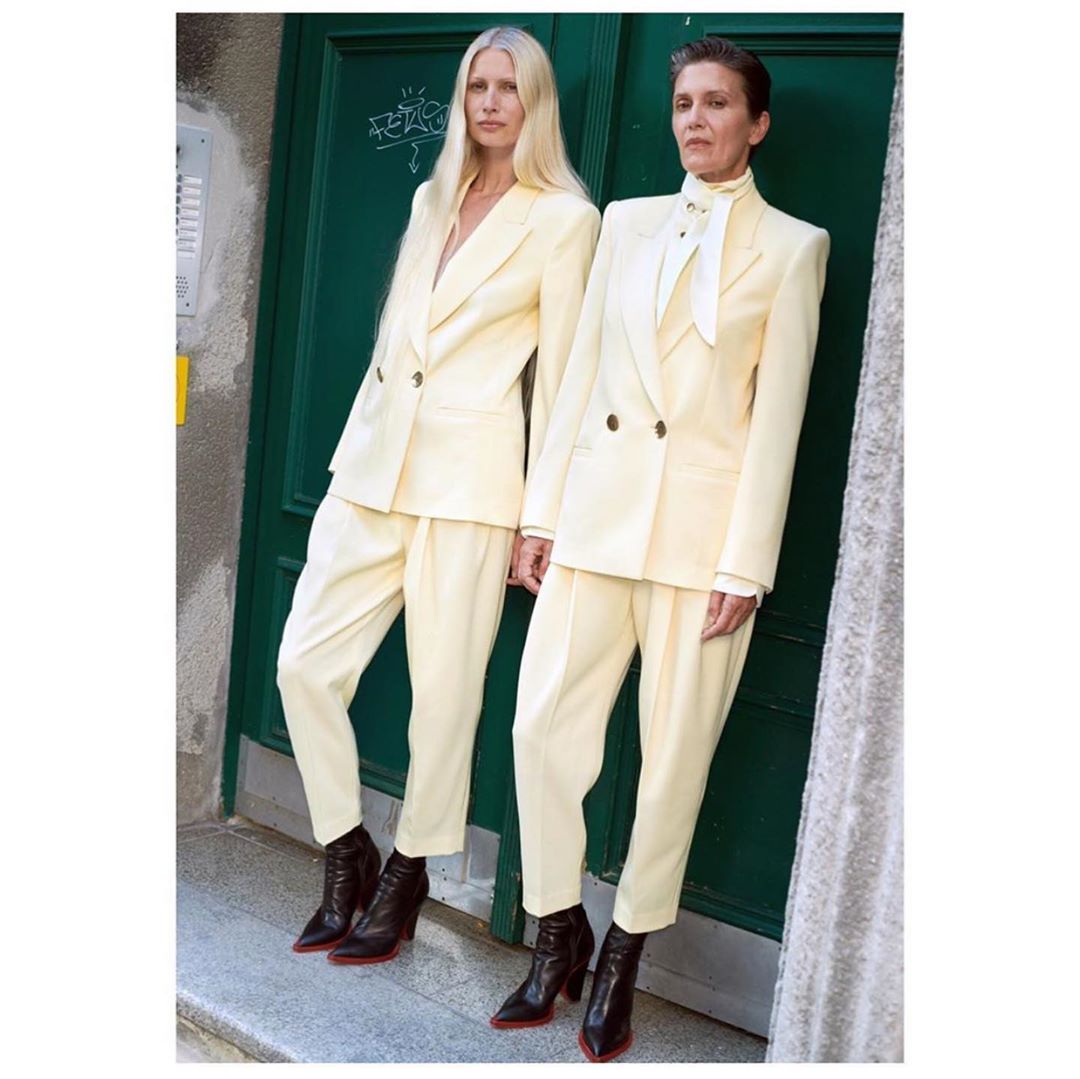 Two women wearing matching cream-colored pant suits pose in front of a green door with dark brown ankle boots with red soles.