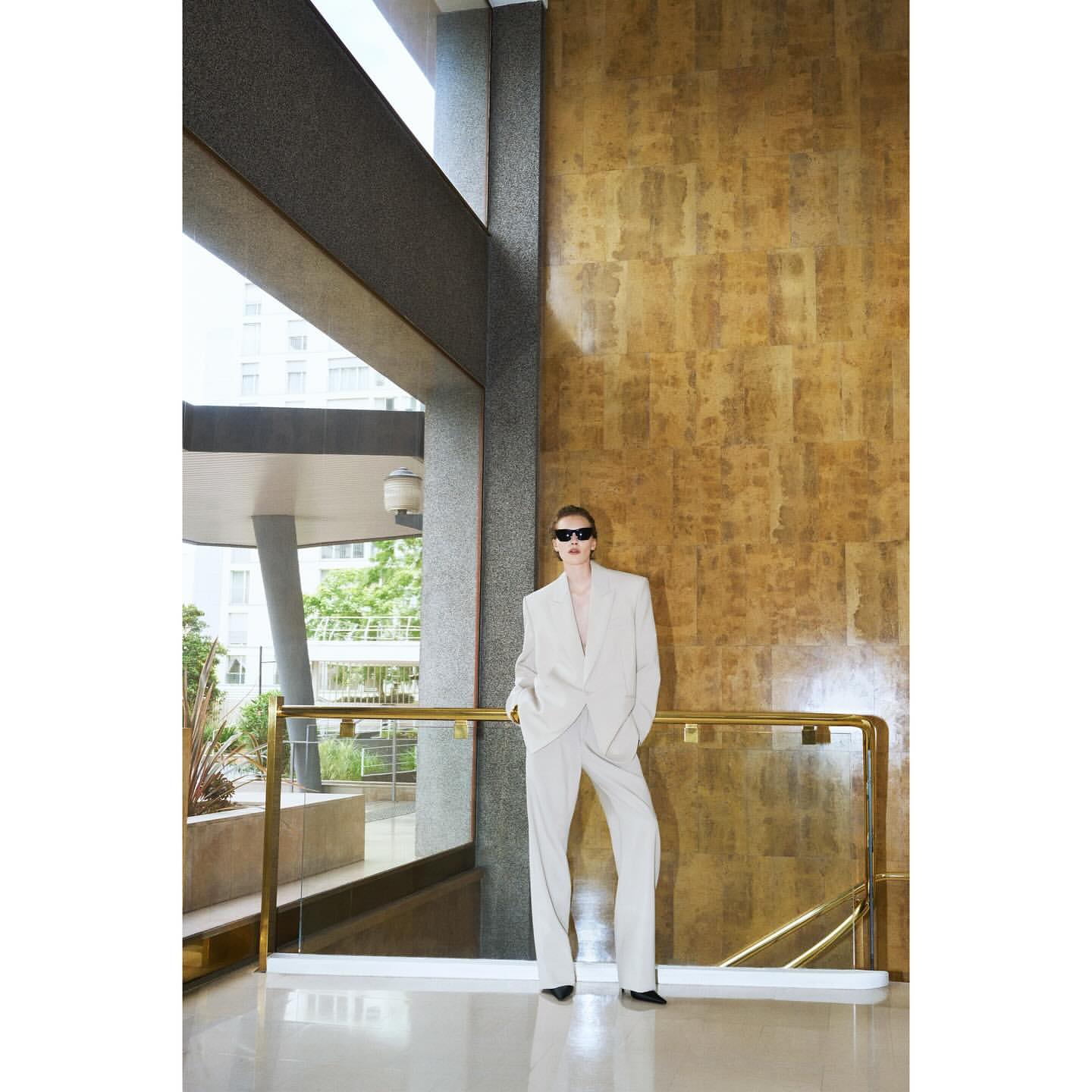 An adult female poses in a cream-colored suit with sunglasses, on a building balcony with gold paneling and modern architecture.