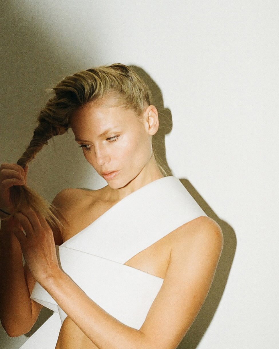 Blonde woman with fair skin poses in a studio, holding her twisted hair and wearing a white sleeveless garment. Beauty shot.