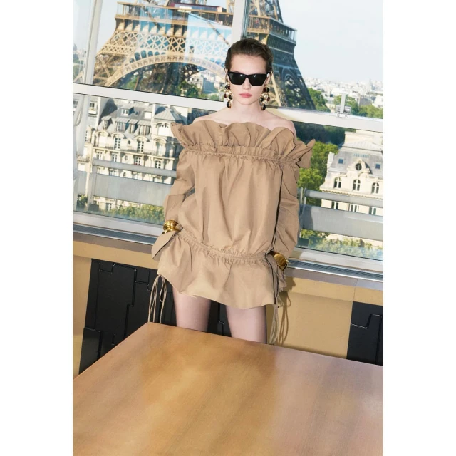 Adult female model poses in a fashionable off-the-shoulder tan dress with the Eiffel Tower visible through a window in the background.
