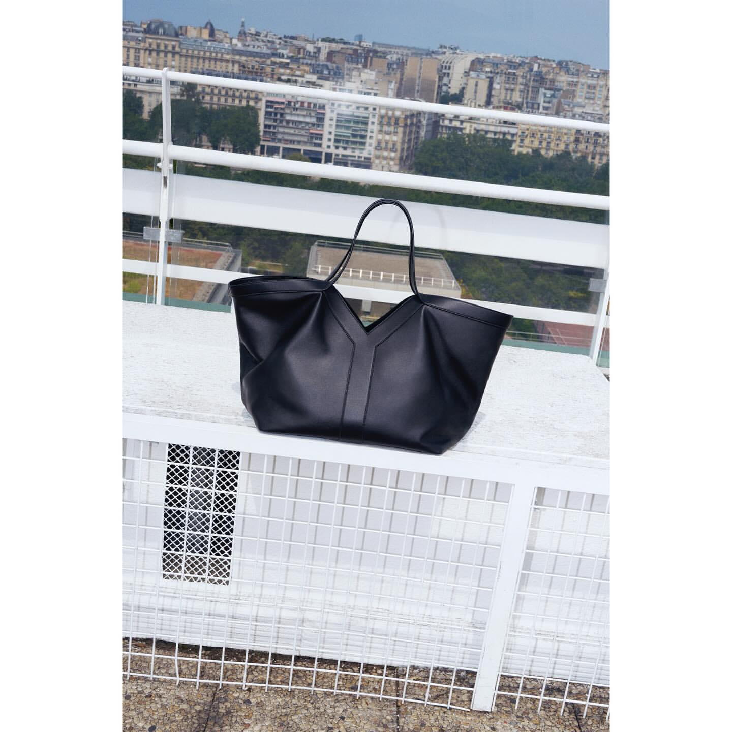 Elegant black leather tote bag with long handles on a white lattice table against an urban cityscape backdrop, minimalist fashion.